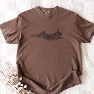 Puede incluir: Una camiseta marr&oacute;n con una ilustraci&oacute;n negra de un conejo corriendo. La camiseta tiene mangas cortas y cuello redondo. La tela tiene un aspecto ligeramente descolorido.