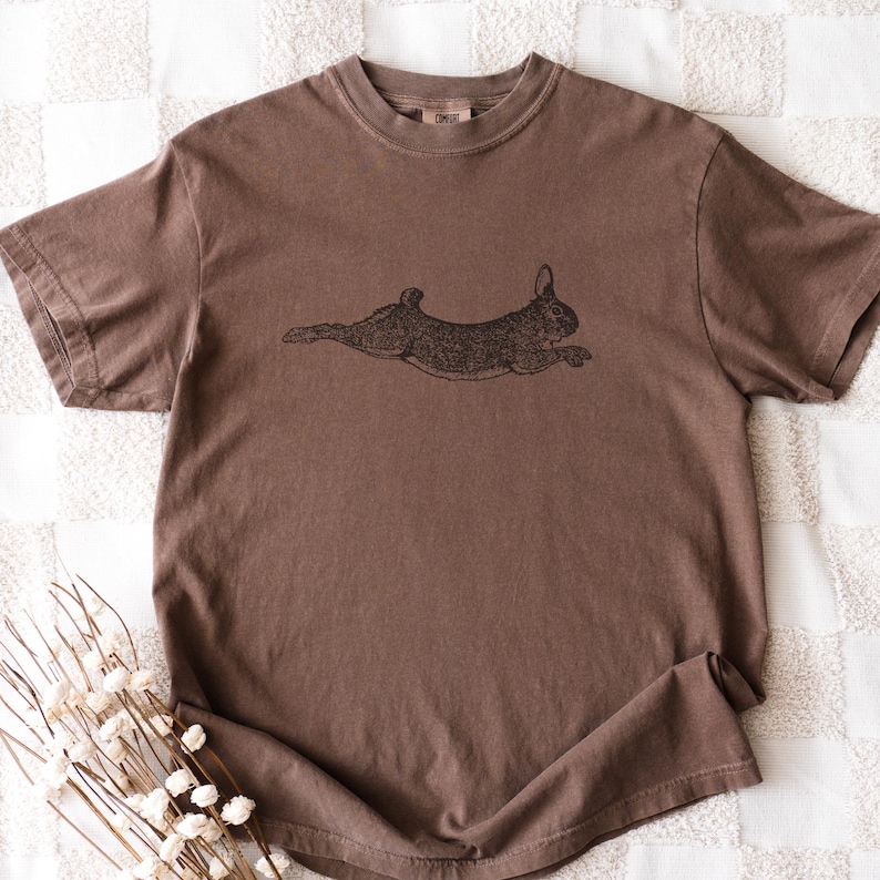 Puede incluir: Una camiseta marr&oacute;n con una ilustraci&oacute;n negra de un conejo corriendo. La camiseta tiene mangas cortas y cuello redondo. La tela tiene un aspecto ligeramente descolorido.