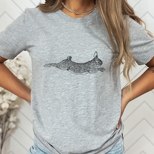 Puede incluir: Una camiseta gris claro con una ilustraci&oacute;n detallada en blanco y negro de un conejo corriendo. El conejo se representa en una pose din&aacute;mica, con las patas extendidas como si estuviera en pleno salto. La camiseta est&aacute; hecha de un material suave.
