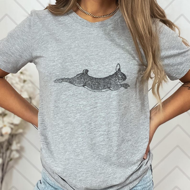 Puede incluir: Una camiseta gris claro con una ilustraci&oacute;n detallada en blanco y negro de un conejo corriendo. El conejo se representa en una pose din&aacute;mica, con las patas extendidas como si estuviera en pleno salto. La camiseta est&aacute; hecha de un material suave.