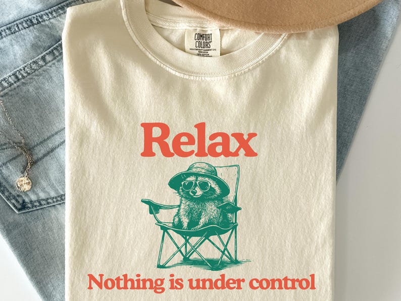 May include: Cream-colored t-shirt with the word "Relax" in orange block letters. Below the word is a graphic of a raccoon wearing sunglasses and a hat, sitting in a folding chair. The phrase "Nothing is under control" is printed below the graphic.