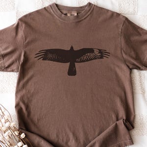 May include: A brown t-shirt with a dark silhouette of a bird in flight. The bird's wings are spread wide, and the design is centered on the shirt. The shirt is made of a soft material and has a classic crew neck.