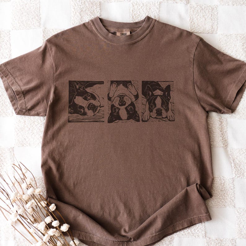May include: Brown t-shirt featuring a graphic of three Boston Terriers in black and white. The dogs are in different poses, with one upside down. The shirt has a crew neck and short sleeves. The fabric has a slightly faded appearance.