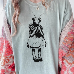 Puede incluir: Camiseta azul claro con una ilustraci&oacute;n en blanco y negro de una mantis religiosa con un vestido. La mantis tiene las manos juntas. El vestido tiene un estampado de lunares. La camiseta es de material suave.