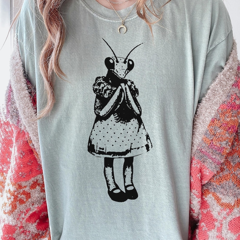 Puede incluir: Camiseta azul claro con una ilustraci&oacute;n en blanco y negro de una mantis religiosa con un vestido. La mantis tiene las manos juntas. El vestido tiene un estampado de lunares. La camiseta es de material suave.