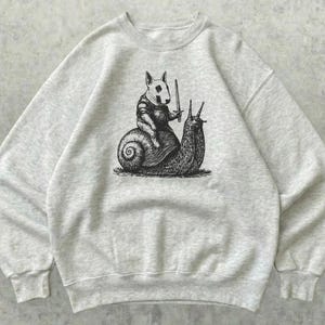 Peut inclure: Un sweat-shirt à col rond gris chiné clair avec une illustration à l'encre noire sur la poitrine. Le dessin représente un bull terrier portant une armure et tenant une épée, chevauchant le dos d'un grand escargot de jardin.