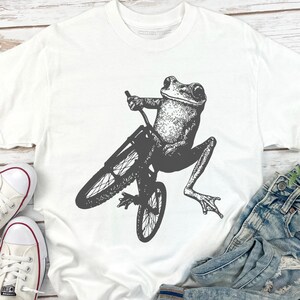 Pu&ograve; includere: T-shirt bianco con un'illustrazione dettagliata in bianco e nero di una rana in bicicletta. La rana &egrave; al centro, con le ruote e il telaio della bicicletta ben visibili. La maglietta &egrave; realizzata in un materiale morbido e confortevole.