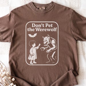 May include: Brown t-shirt with a white graphic that reads "Don't Pet the Werewolf." The graphic depicts a child reaching toward a werewolf, with a bat above them. The shirt is made of a soft material.