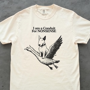 May include: Cream-colored t-shirt with a black graphic of a dog riding a goose with wings. The text "I am a Conduit For NONSENSE" is above the illustration. The shirt has a crew neck and short sleeves.
