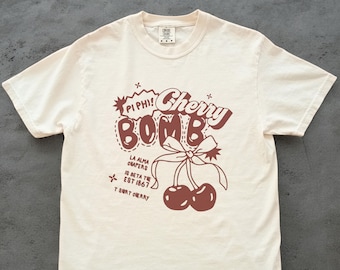 May include: Cream-colored t-shirt featuring a brown graphic design. The design includes the words "Cherry Bomb" in a playful font, along with the text "Pi Phi!" and "La Alma Chapers". Two cherries with a bow are also depicted.