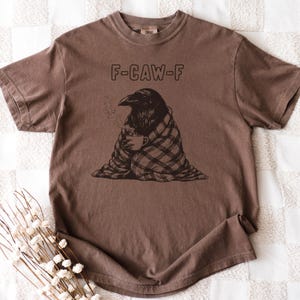 May include: Brown t-shirt with the text "F-CAW-F" above a graphic of a raven wrapped in a plaid blanket, holding a cup of coffee. The raven is depicted with steam rising from the cup. The shirt has short sleeves and a crew neck.