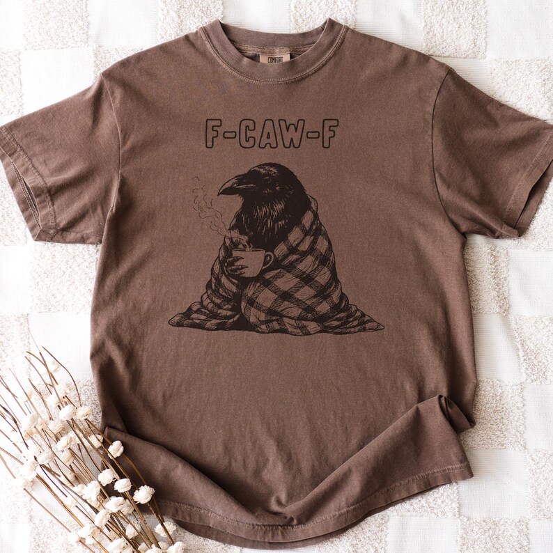 May include: Brown t-shirt with the text "F-CAW-F" above a graphic of a raven wrapped in a plaid blanket, holding a cup of coffee. The raven is depicted with steam rising from the cup. The shirt has short sleeves and a crew neck.
