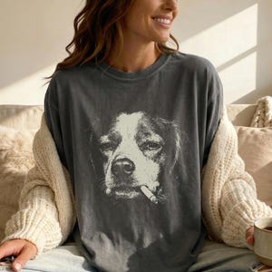 Könnte beinhalten: Dunkelgraues T-Shirt mit einem schwarz-weißen Hund, der eine Zigarette im Maul hat. Das Gesicht des Hundes ist das zentrale Bild, mit detailliertem Fell und Ausdruck. Das T-Shirt hat eine lockere Passform.