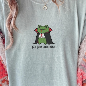 May include: A pale blue t-shirt with a cartoon frog dressed as a vampire, wearing a black cape and red details. The text "pls just one bite" is below the frog. The shirt is a casual style.