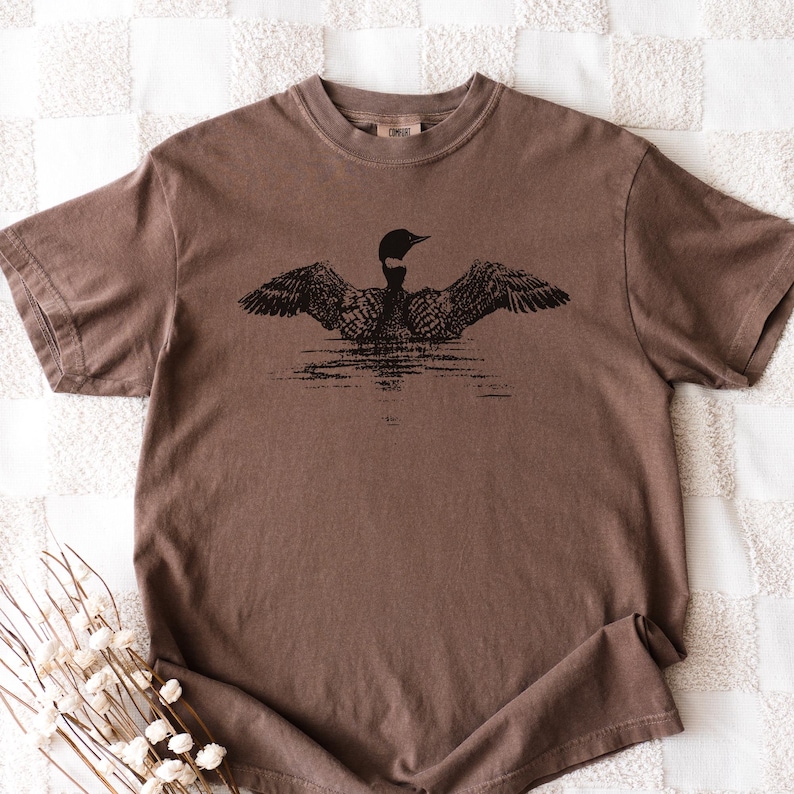 May include: A brown t-shirt featuring a black illustration of a loon with outstretched wings, appearing to float on water. The shirt has a crew neck and short sleeves. The fabric has a slightly faded appearance.