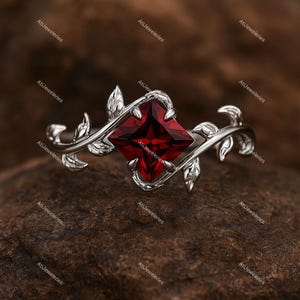 May include: A silver ring featuring a square, deep red gemstone. The ring's band is adorned with a delicate, vine-like design with leaves. The gemstone is the focal point, set in a prong setting, and the ring is displayed on a textured surface.