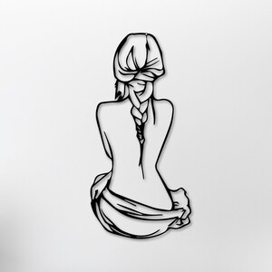 May include: Black metal wall art sculpture of a woman sitting with her back to the viewer, her hair in a braid. The sculpture is mounted on a white wall in a living room setting.