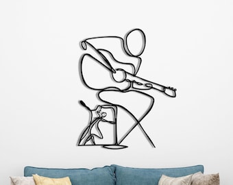 May include: Black metal wall art depicting a musician playing a guitar. The artwork features a minimalist, continuous line design of a person seated on a stool, with a guitar. The art is mounted on a white wall above a blue sofa.