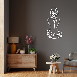 May include: White metal wall art depicting a woman sitting with her back to the viewer, her hair in a braid, and a flowing garment draped around her.