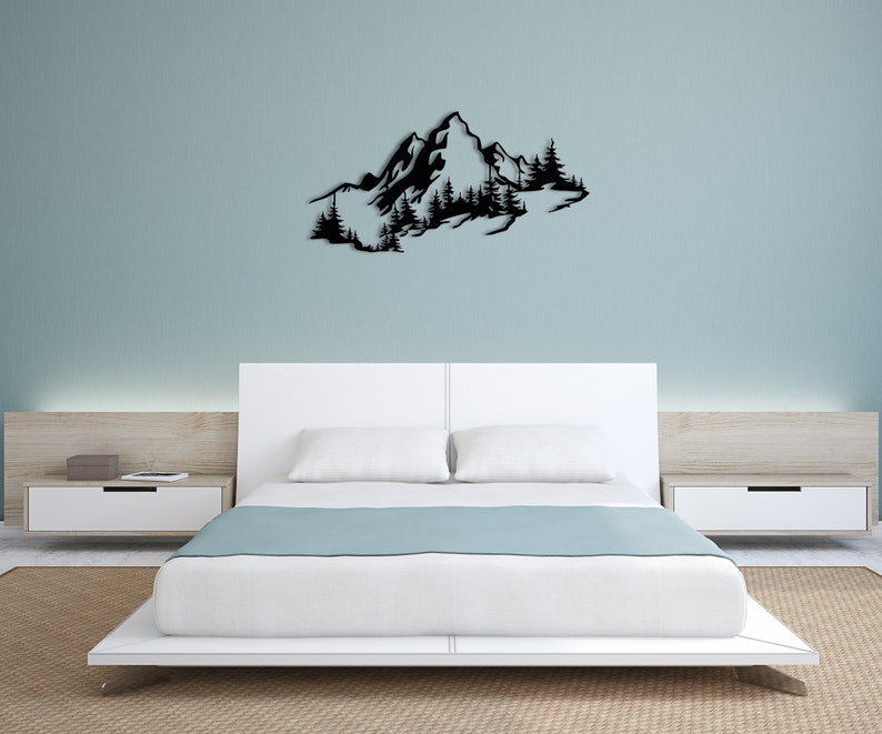 May include: Black metal wall art depicting a mountain range with trees. The art is mounted on a white wall above a white bed with a light blue blanket.