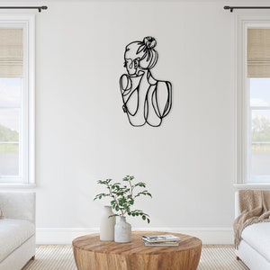 May include: Black metal wall art sculpture of a woman with her hair in a bun. The sculpture is in a minimalist style and is a modern home decor piece.
