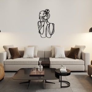 May include: A black metal wall art sculpture depicting a stylized outline of a woman with her hair pulled back in a bun. The sculpture is hanging on a white wall in a living room setting with a couch, coffee table, and floor lamp.