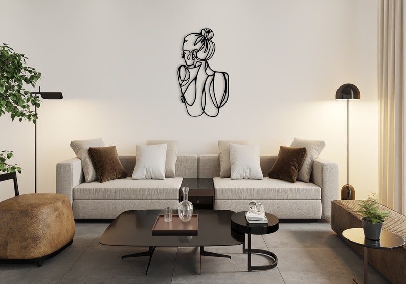 May include: A black metal wall art sculpture depicting a stylized outline of a woman with her hair pulled back in a bun. The sculpture is hanging on a white wall in a living room setting with a couch, coffee table, and floor lamp.