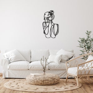 May include: A white sofa with two white pillows and a wicker chair with a white cushion. A small, round, woven ottoman sits in front of the sofa. A black metal wall sculpture of a woman's profile is on the wall behind the sofa. A potted plant sits on a wooden table to the left of the sofa. A potted plant sits on the floor to the right of the chair.