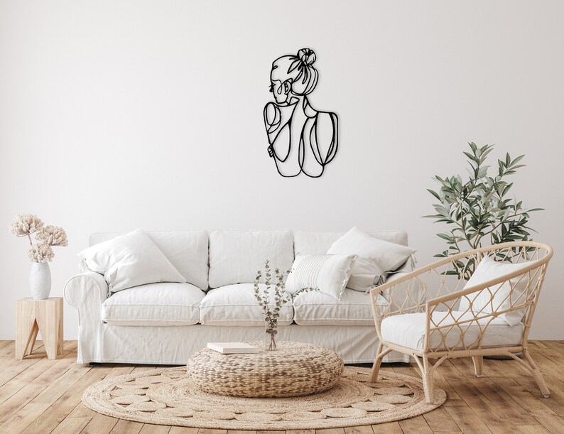 May include: A white sofa with two white pillows and a wicker chair with a white cushion. A small, round, woven ottoman sits in front of the sofa. A black metal wall sculpture of a woman's profile is on the wall behind the sofa. A potted plant sits on a wooden table to the left of the sofa. A potted plant sits on the floor to the right of the chair.