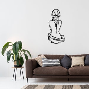 May include: Black metal wall art depicting a woman sitting with her back to the viewer, her hair in a braid. The art is mounted on a white wall above a brown couch with throw pillows.