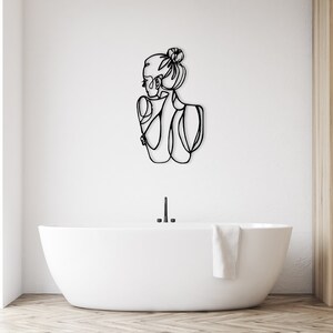 May include: A black line art wall decor of a woman with her hair in a bun, standing in a bathroom with a white bathtub and a wooden shelf unit.