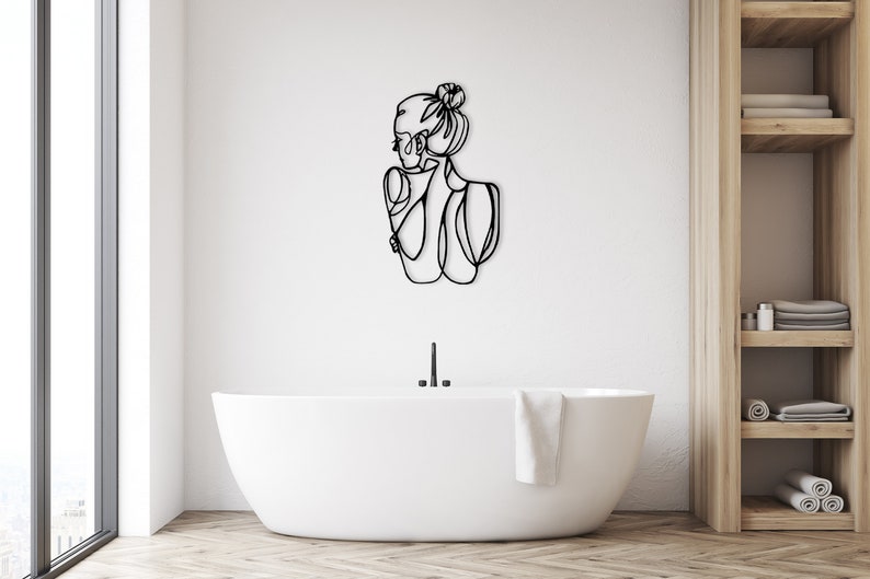 May include: A black line art wall decor of a woman with her hair in a bun, standing in a bathroom with a white bathtub and a wooden shelf unit.