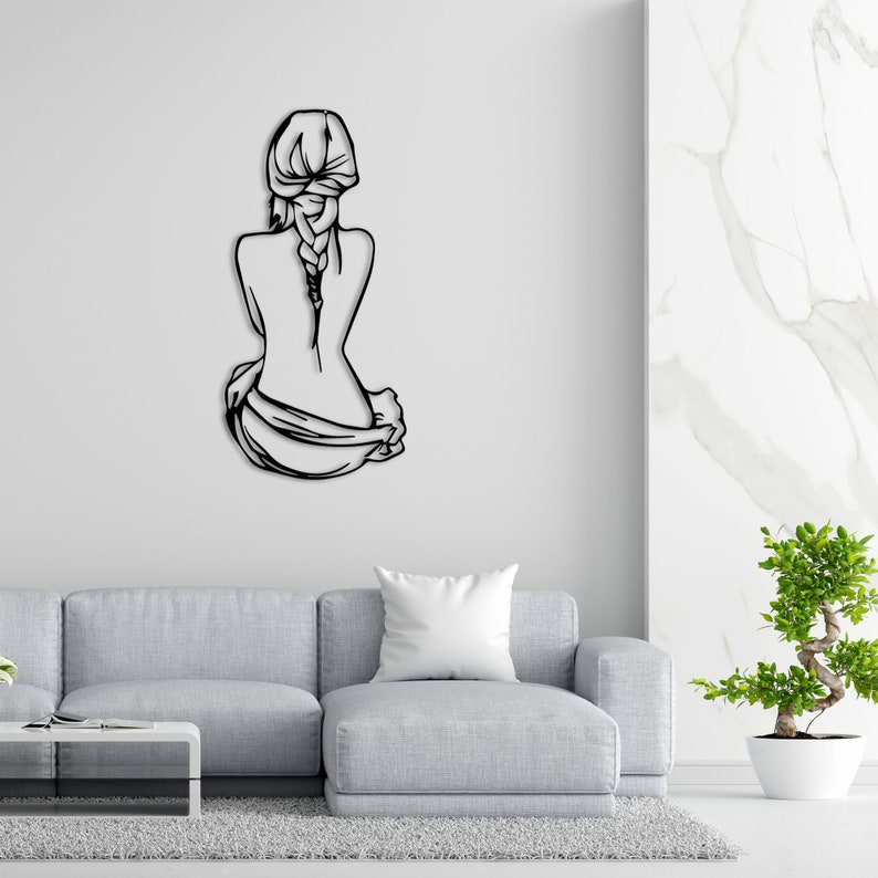 May include: Black metal wall art depicting a woman sitting with her back to the viewer, her hair in a braid, and a cloth draped over her legs.