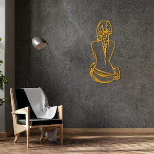 May include: A gold metal wall decor featuring a stylized outline of a person sitting with their back to the viewer. The person has long hair and is wearing a flowing garment.