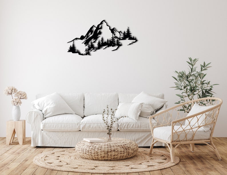 May include: Black metal wall art depicting a mountain range with trees. The art is displayed on a white wall above a white couch and a wicker chair.