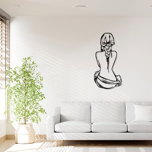 May include: Black metal wall art depicting a woman sitting with her back to the viewer, her hair in a braid. The art is mounted on a white wall in a living room setting.