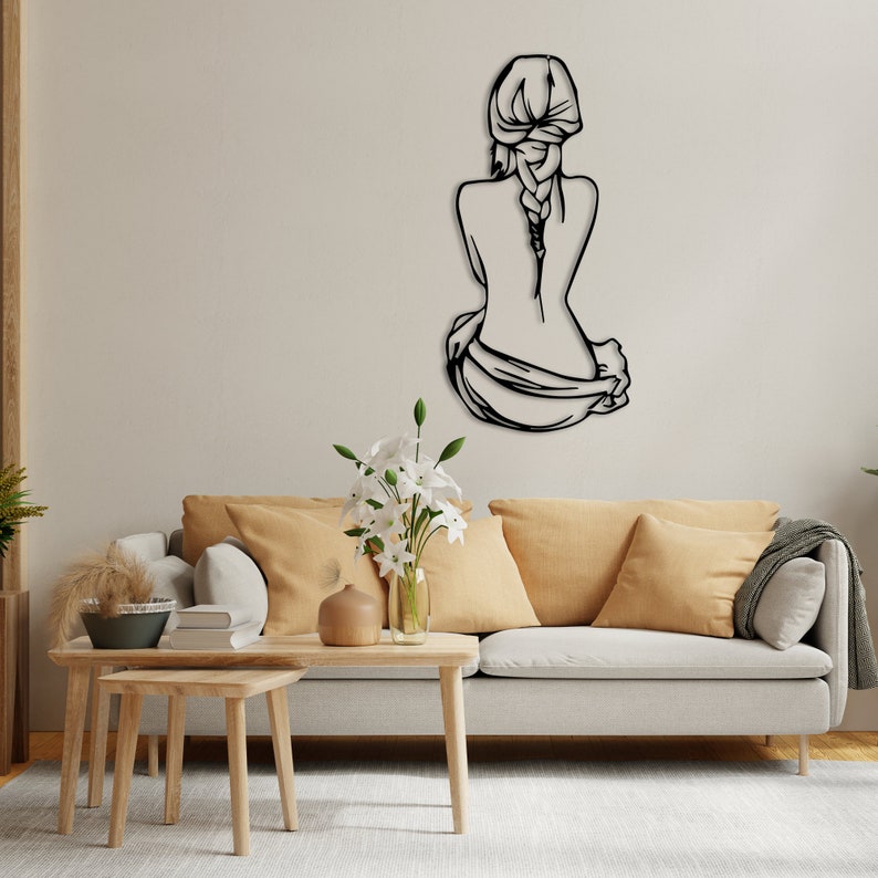 May include: Black metal wall art depicting a woman sitting with her back to the viewer, her hair in a braid. The art is mounted on a white wall above a light gray couch with beige throw pillows.