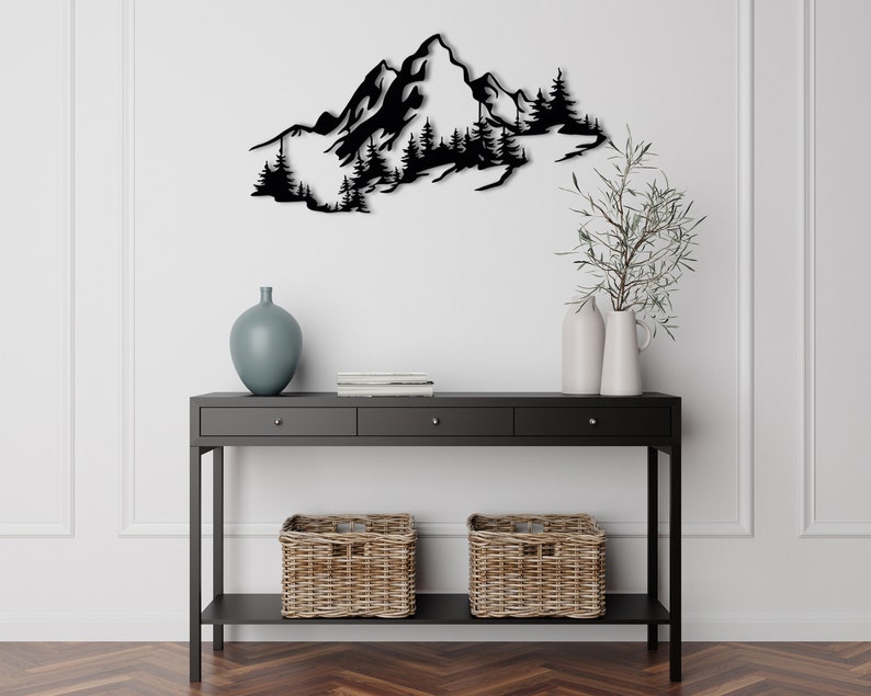 May include: Black metal wall art depicting a mountain range with trees. The art is mounted on a white wall above a black console table with two wicker baskets.