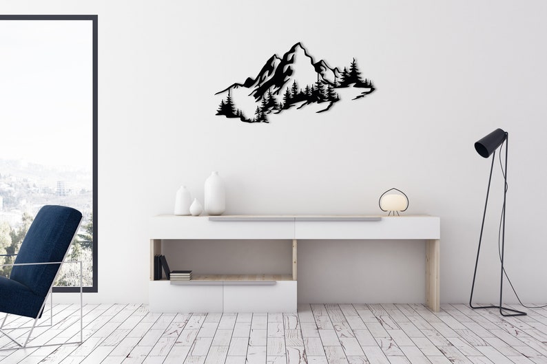 May include: Black metal wall art depicting a mountain range with trees. The art is mounted on a white wall above a white shelf with a lamp and decorative items.