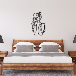 May include: A black metal wall art sculpture of a woman with her hair in a bun. The sculpture is hanging on a white wall above a wooden bed frame with gray and white pillows.