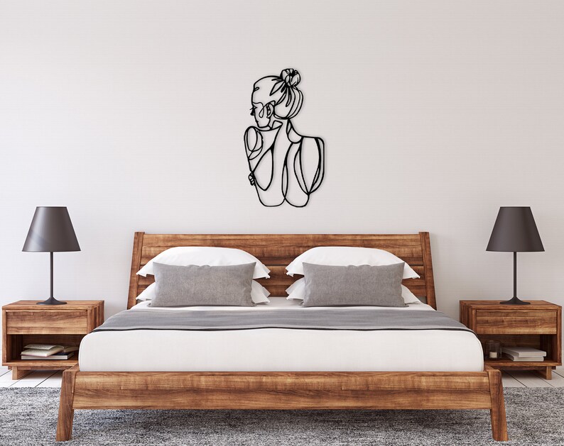 May include: A black metal wall art sculpture of a woman with her hair in a bun. The sculpture is hanging on a white wall above a wooden bed frame with gray and white pillows.