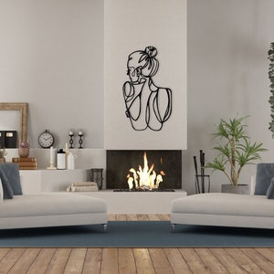 May include: A black metal wall sculpture of a woman's profile with a bun in her hair. The sculpture is hanging on a white wall above a fireplace with a fire burning inside. There are two white couches in the room, one on each side of the fireplace.
