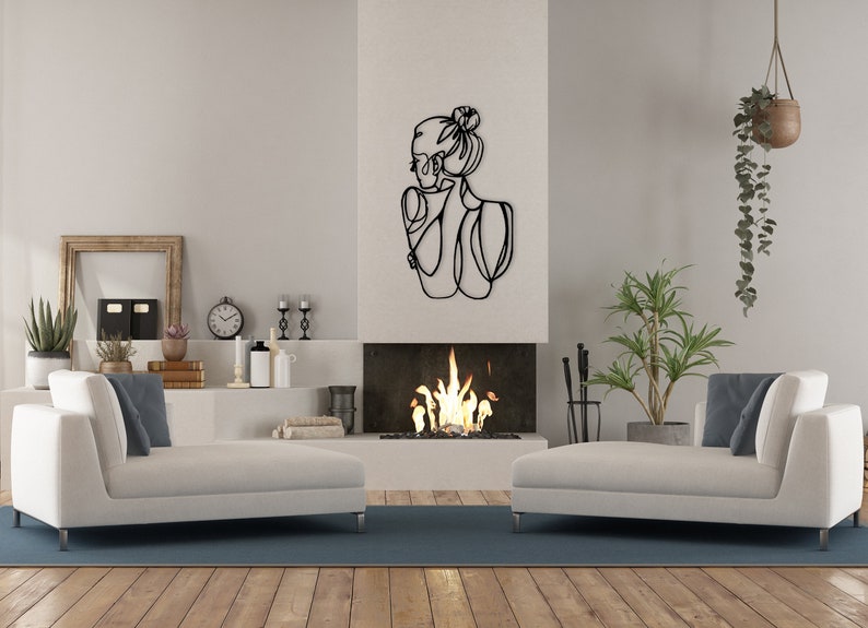 May include: A black metal wall sculpture of a woman's profile with a bun in her hair. The sculpture is hanging on a white wall above a fireplace with a fire burning inside. There are two white couches in the room, one on each side of the fireplace.