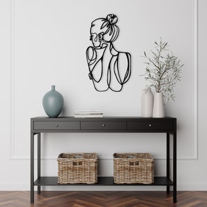 May include: Black metal wall art depicting a woman's silhouette with a bun in her hair. The art is mounted on a white wall above a black console table with two drawers and a lower shelf. The table is decorated with two wicker baskets and a blue vase.
