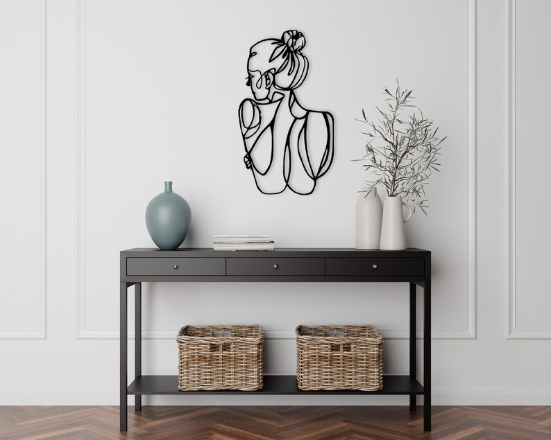 May include: Black metal wall art depicting a woman's silhouette with a bun in her hair. The art is mounted on a white wall above a black console table with two drawers and a lower shelf. The table is decorated with two wicker baskets and a blue vase.