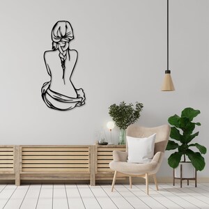 May include: Black metal wall art depicting a woman sitting with her back to the viewer, her hair in a braid. The art is mounted on a white wall in a room with a wooden cabinet, a chair, and plants.