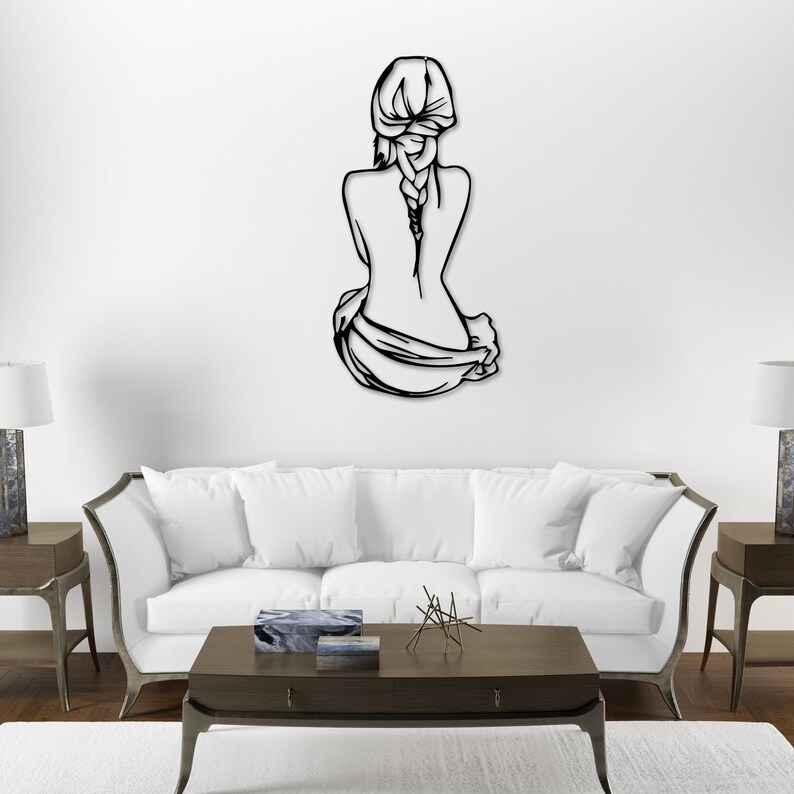 May include: Black metal wall art sculpture of a woman sitting with her back to the viewer, her hair in a braid. The sculpture is mounted on a white wall in a living room setting.