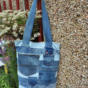 May include: A tote bag constructed from horizontal strips of light and medium blue denim fabric. The bag features multiple exterior pockets salvaged from jeans, including two large patch pockets and two smaller coin pockets, with two long shoulder straps.