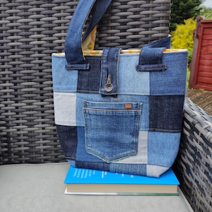 May include: A patchwork denim tote bag with a front pocket and button closure. The bag features various shades of blue denim squares and a denim handle. The bag is sitting on a blue book.