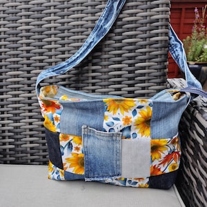 May include: A patchwork denim shoulder bag with a floral sunflower pattern. The bag has a blue denim strap and a front pocket. The bag is made of various denim patches and floral fabric.